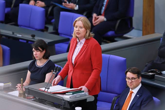 Bärbel Bas in a red jacket standing at the President’s desk and speaking, with a parliamentary secretary seated beside her