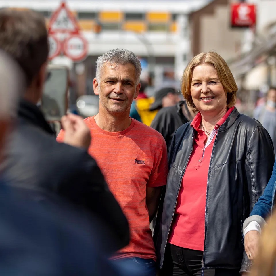 Bärbel Bas, smiling in a black leather jacket and pink shirt, standing on a busy shopping street next to a man in an orange T-shirt