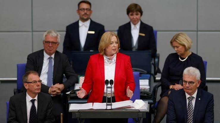 Bärbel Bas in a red jacket speaking from the President’s desk in the Bundestag plenary chamber, with officials seated behind her