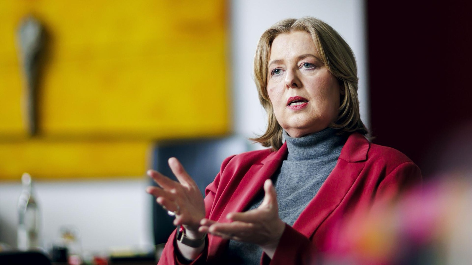 Bärbel Bas in a red blazer and grey roll-neck, gesturing with both hands while speaking during an interview