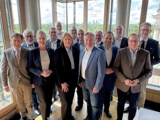 Group photograph of about fourteen people in business attire, including Bärbel Bas in a dark suit, standing in front of tall windows with a view over trees and a city skyline